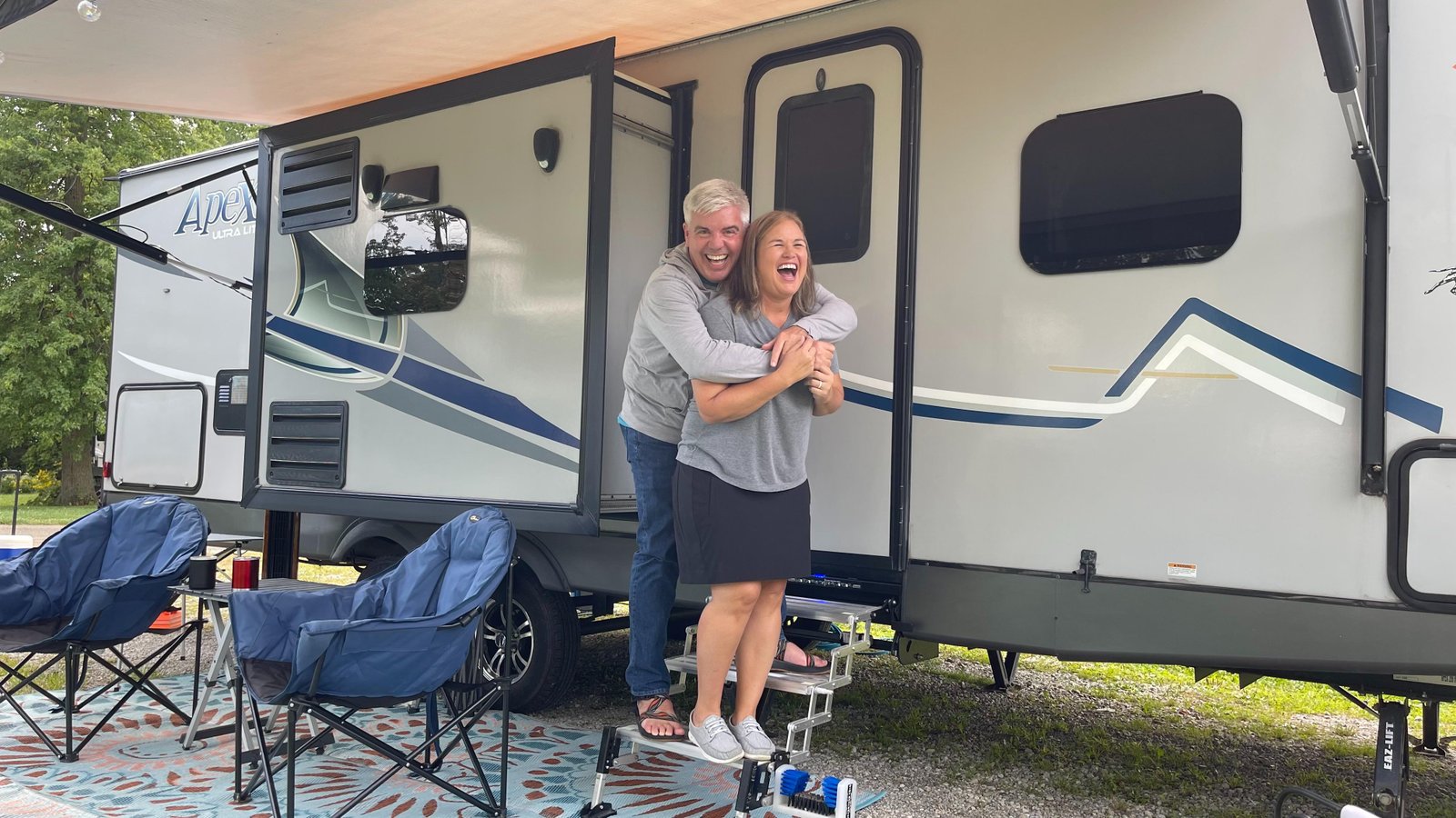 Brian and Michelle laughing in a hug on the steps of a Coachmen Apex travel trailer at a campsite